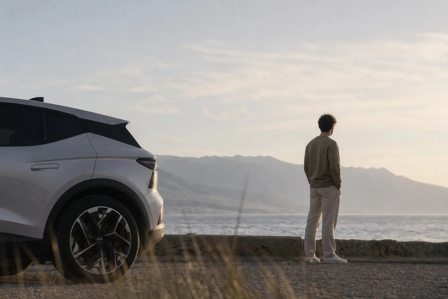 Homme au bord de la mer face à la montagne, à côté d’une Tesla blanche